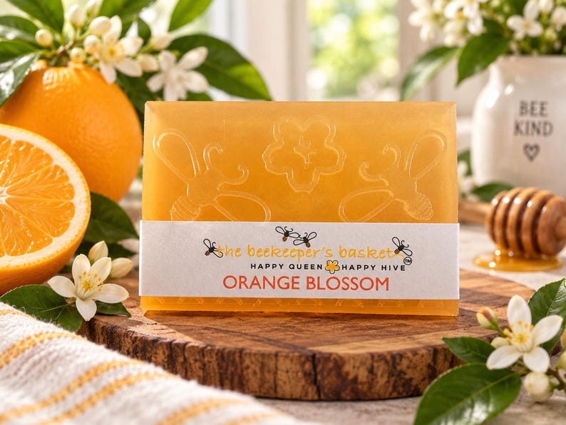 May include: A rectangular orange soap bar embossed with two bees and a flower. A white paper label wraps around the center, printed with the text "the beekeeper's basket", "HAPPY QUEEN HAPPY HIVE", and "ORANGE BLOSSOM" in orange and black ink.
