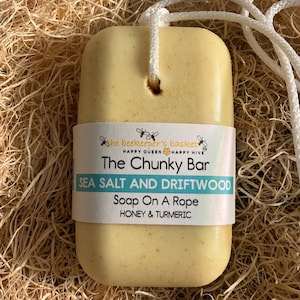 May include: A rectangular, light yellow bar of soap on a rope, with a white label that reads "The Chunky Bar" and "Sea Salt and Driftwood." The soap has a hole for the rope and is packaged on a bed of straw.