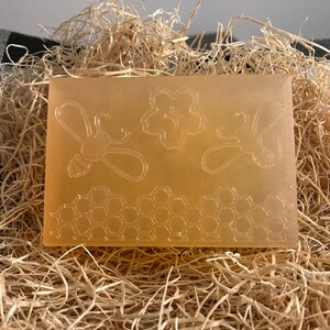May include: A rectangular, light amber soap bar featuring embossed bee and honeycomb designs. The soap is displayed on a bed of straw, suggesting a natural, handcrafted product. The design includes two bees, a flower, and a honeycomb pattern.