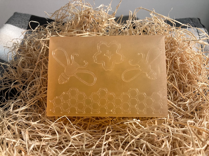 May include: A rectangular, light amber soap bar featuring embossed bee and honeycomb designs. The soap is displayed on a bed of straw, suggesting a natural, handcrafted product. The design includes two bees, a flower, and a honeycomb pattern.