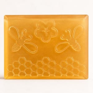 May include: A rectangular, translucent amber-colored bar of soap featuring embossed designs of two bees, a central flower, and a honeycomb pattern along the bottom edge. The soap has a smooth, polished finish and a consistent golden-yellow hue.