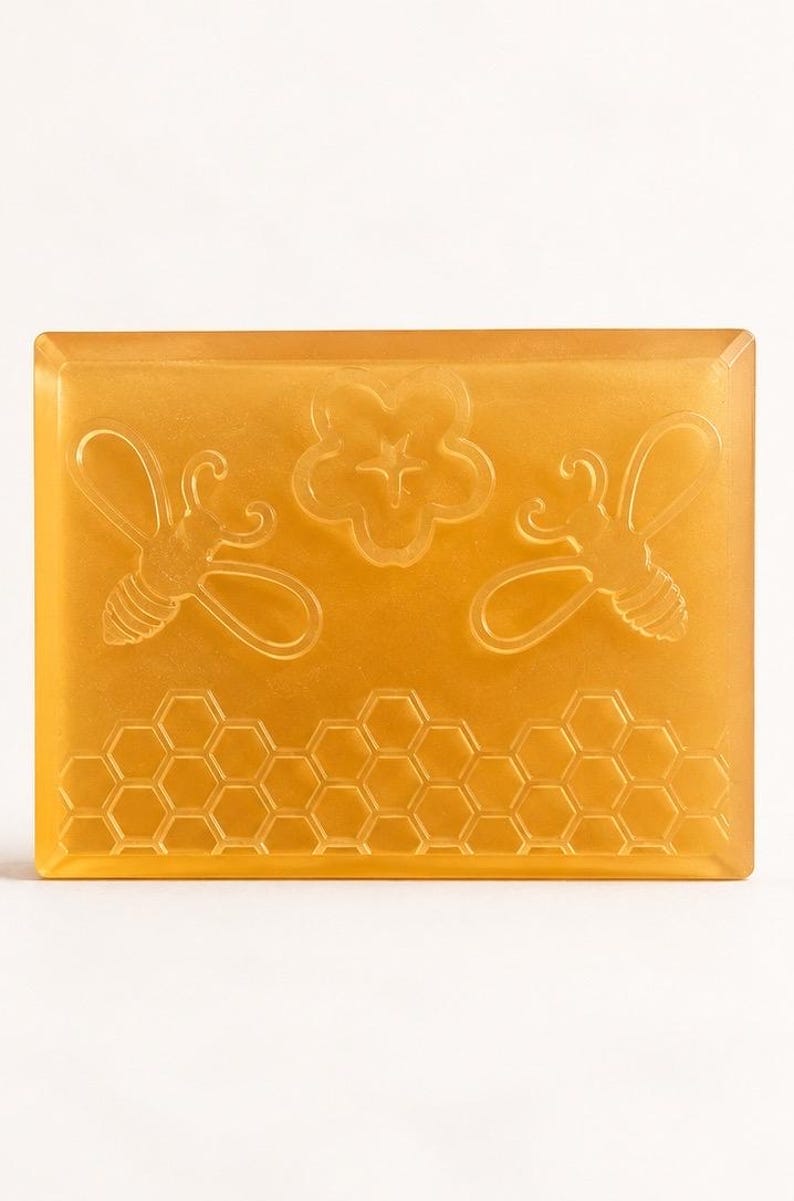 May include: A rectangular, translucent amber-colored bar of soap featuring embossed designs of two bees, a central flower, and a honeycomb pattern along the bottom edge. The soap has a smooth, polished finish and a consistent golden-yellow hue.