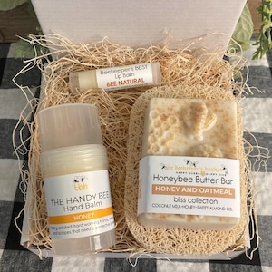 May include: A gift box containing a lip balm, hand balm, and a honey and oatmeal butter bar. The lip balm is labeled "BEE NATURAL". The hand balm is labeled "THE HANDY BEE Hand Balm HONEY". The butter bar is labeled "Honeybee Butter Bar HONEY AND OATMEAL".