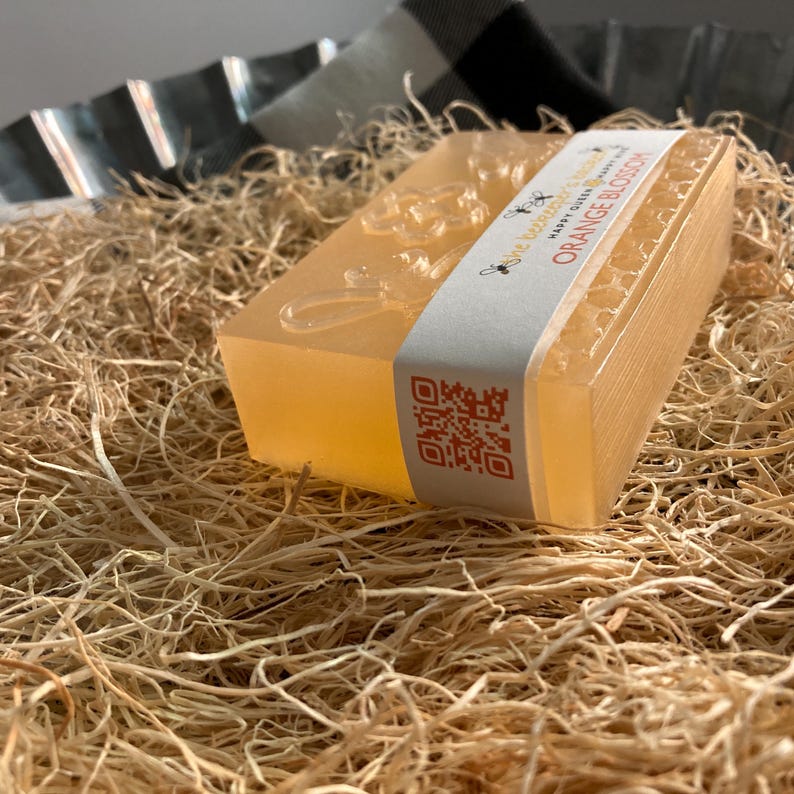 May include: A rectangular bar of orange blossom soap with a white label that reads "Happy Queen Bee" and "Orange Blossom." The soap has a floral design and a QR code. The soap is resting on a bed of straw.