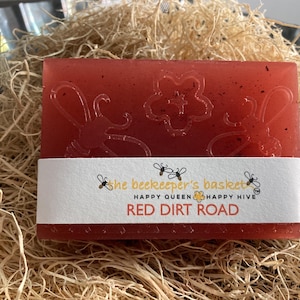 May include: A red bar of soap with a floral design and the text "the beekeeper's basket" "HAPPY QUEEN" "HAPPY HIVE" "RED DIRT ROAD" printed on the label.