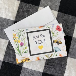 May include: A floral greeting card with the text "just for YOU!" and a yellow heart. The card features illustrations of bees, flowers, and greenery, and comes with a white envelope.