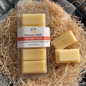 May include: A package of beeswax melts in a clear plastic container. The label reads "tbb Beeswax Melts Red Dirt Road pure beeswax-small batch-hand poured net wt. 1.75 oz."