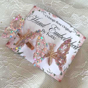 May include: Two pink butterfly hair clips with gold-colored metal accents and glitter. The butterfly wings are embellished with iridescent glitter and star-shaped sequins. The clips are displayed on a card that reads "Hand Embellished By Katie."