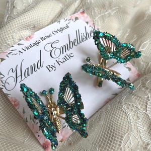 May include: Two handcrafted butterfly hair clips with emerald green glitter wings and gold-colored metal bodies. The clips are displayed on a floral card that reads "Hand Embellished By Katie."