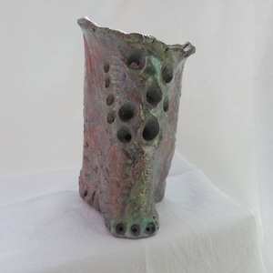 May include: A ceramic vase with a textured, green, brown, and red surface. The vase has a unique, irregular shape with multiple holes throughout the surface.