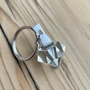 May include: A clear, faceted, diamond-shaped keyring with a silver key ring. The keyring is attached to a silver metal loop.
