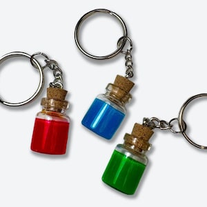 May include: Three glass bottle keychains with cork stoppers, each filled with a different colored liquid: red, blue, and green. The bottles are attached to silver key rings via small chains. The background is white.
