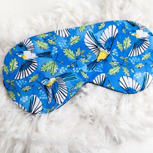 May include: A blue sleep mask with a pattern of birds, leaves, and berries. The sleep mask is oval-shaped with a black border. The background is white, fluffy material.