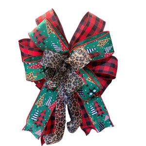 May include: A large Christmas bow made with three different ribbons. The ribbons are red and black buffalo plaid, green with white and black Christmas trees, and leopard print.