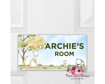 May include: A rectangular sign with the text "ARCHIE'S ROOM" in green. The sign features a watercolour illustration of a kangaroo, a tree, and other animals. The sign is decorated with a floral arrangement and the text "Paper Shed Designs Stationery & Gifts".