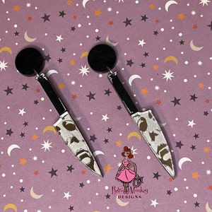 May include: A pair of black and white knife earrings with a spooky face design. The earrings are on a purple background with white stars and moons.