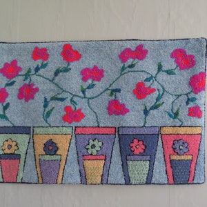 May include: A blue rug featuring a design of pink flowers in pots. The pots are arranged in a row and have different coloured details.