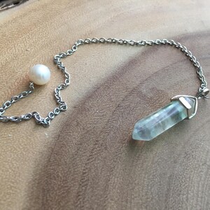 Puede incluir: Una cadena de plata con un péndulo de cristal de fluorita verde y una perla blanca en la parte superior.