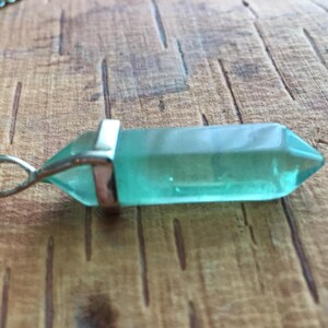 May include: A silver chain necklace with a light blue crystal pendant. The pendant is shaped like a long, thin point.