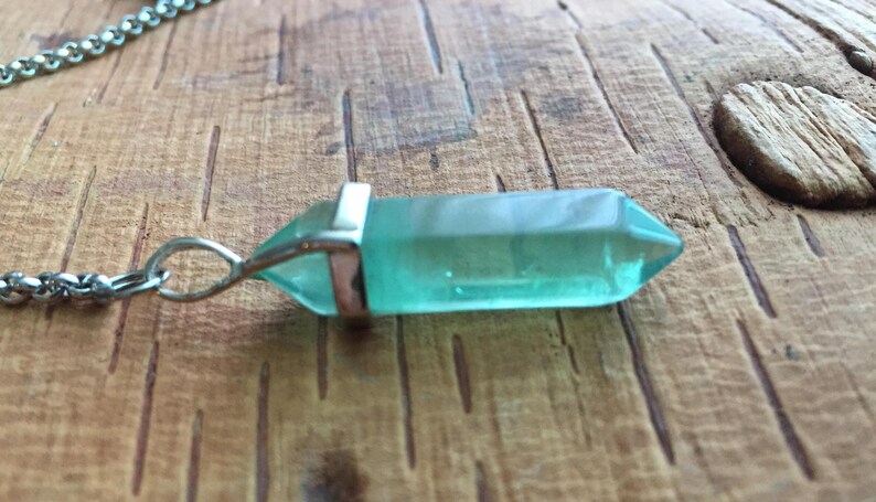 May include: A silver chain necklace with a light blue crystal pendant. The pendant is shaped like a long, thin point.