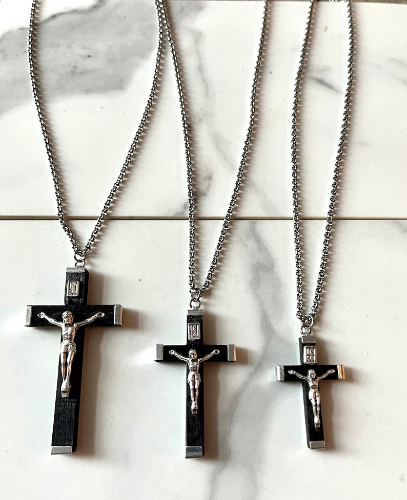 May include: Three silver chain necklaces with black wooden crucifix pendants. The crucifixes feature a silver Jesus figure and are different sizes.