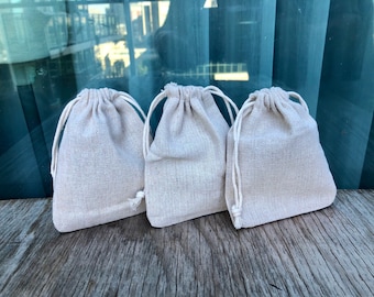 May include: Three small, off-white, drawstring fabric bags made of natural linen. The bags are sitting on a wooden surface.
