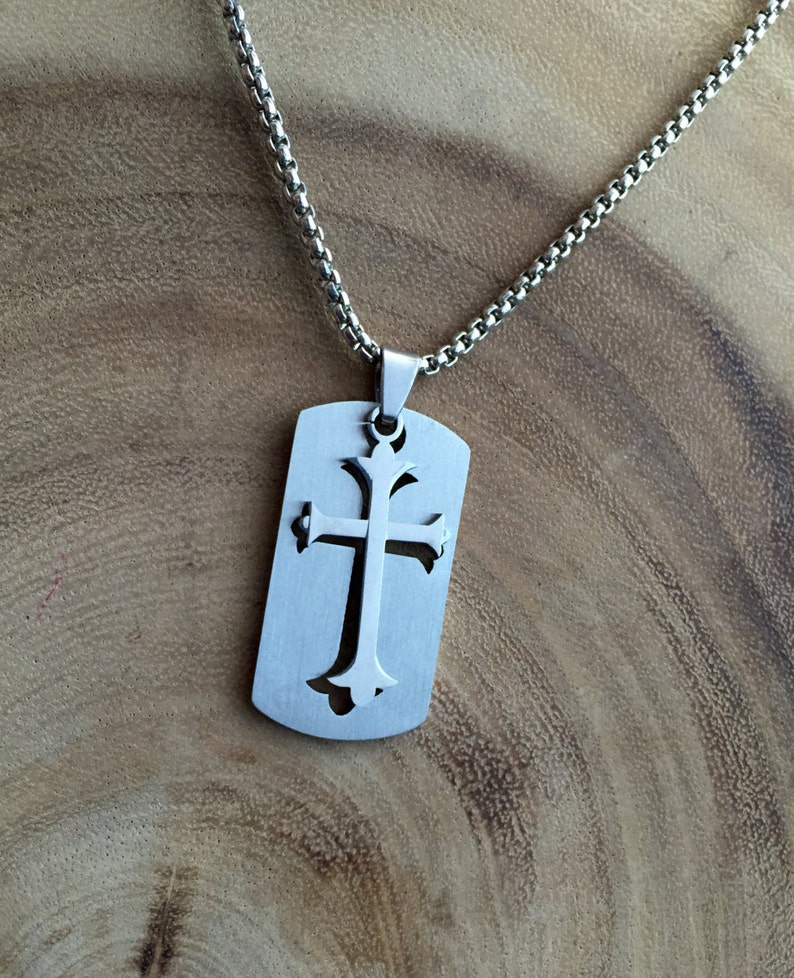 K&ouml;nnte beinhalten: Eine silberne Kettenkette mit einem silbernen Anh&auml;nger in Form eines Hundemarken. Die Hundemarke hat ein ausgeschnittenes Design eines Kreuzes.
