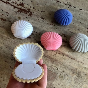 May include: Five small seashell-shaped boxes in different colors. The boxes are made of velvet and have a soft, plush texture. The boxes are open and empty, ready to be filled with jewelry or other small treasures.