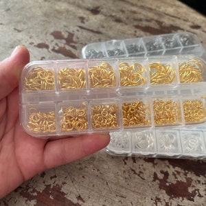 May include: Two clear plastic containers filled with gold jump rings. The containers are divided into multiple compartments.