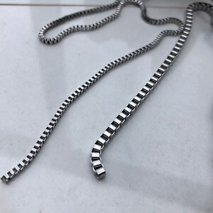 May include: A silver chain necklace with a box chain design. The chain is lying flat on a white surface.