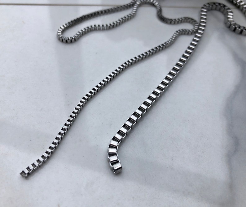 May include: A silver chain necklace with a box chain design. The chain is lying flat on a white surface.
