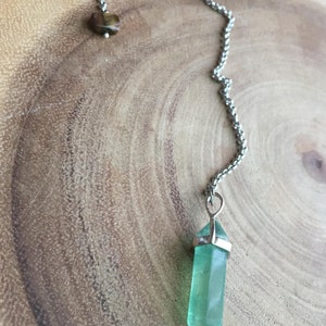 May include: A green crystal pendulum with a silver chain and a brown tiger's eye stone at the top.