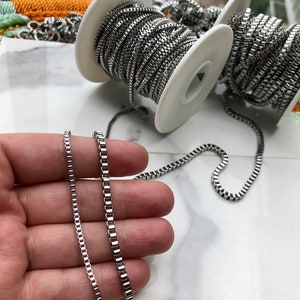 May include: Two silver chains of different lengths, one is longer than the other, both are on a hand, the chains are on spools in the background.