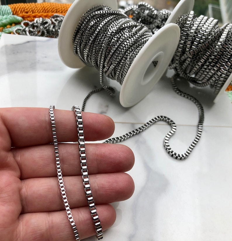 May include: Two silver chains of different lengths, one is longer than the other, both are on a hand, the chains are on spools in the background.