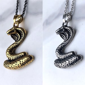 Op de afbeelding: Twee slangen hangerkettingen, een gouden en een zilveren, met een gedetailleerd cobra ontwerp. De kettingen worden getoond op een witte achtergrond.