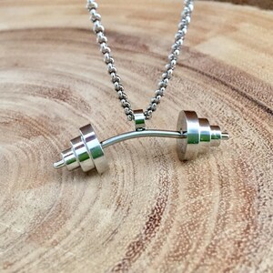 May include: A silver necklace with a barbell pendant. The barbell is made of metal and has a curved shape. The necklace is on a wooden surface.