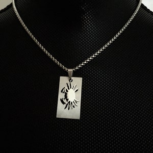 May include: A silver chain necklace with a pendant featuring a cutout design of a crab. The pendant is rectangular and hangs from a silver chain.