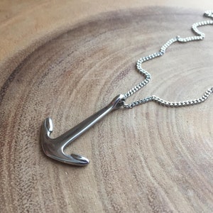 Op de afbeelding: Een zilverkleurige ankerhanger ketting. De ankerhanger heeft een gebogen haak en een rechte schacht. De ketting is een delicate, geschakelde stijl. De ketting wordt gepresenteerd op een houten oppervlak.