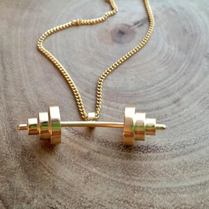 May include: A gold chain necklace with a gold barbell pendant. The barbell is made of multiple stacked rings.