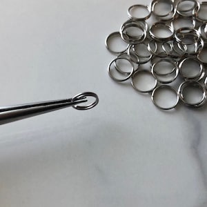 Puede incluir: Primer plano de un anillo de salto plateado sostenido por unas pinzas. En el fondo hay una pila de anillos de salto plateados.