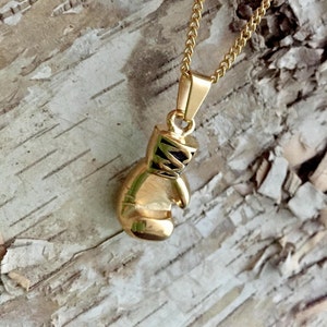May include: A gold-colored boxing glove pendant necklace. The pendant is detailed with black stitching and a black strap. The necklace is on a gold chain.