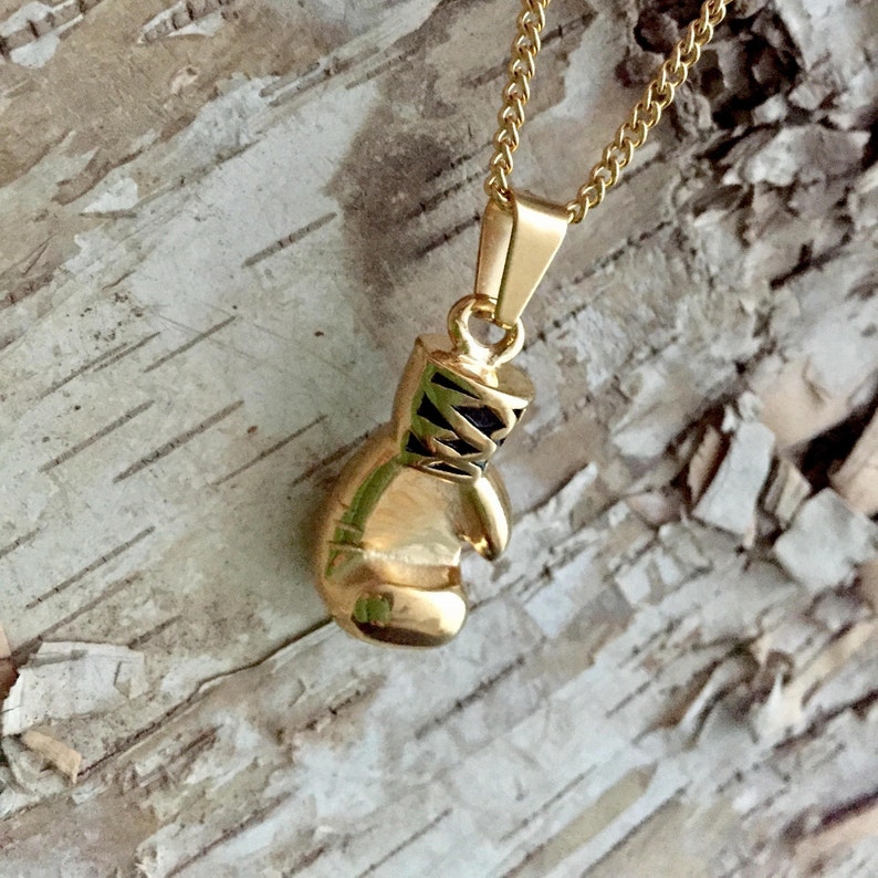May include: A gold-colored boxing glove pendant necklace. The pendant is detailed with black stitching and a black strap. The necklace is on a gold chain.