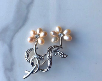May include: Silver brooch with two pearl flowers. Each flower has a small rhinestone in the center. The flowers are connected by a silver stem.