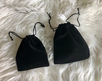 May include: Two black velvet drawstring pouches, one larger than the other, on a white fluffy surface.