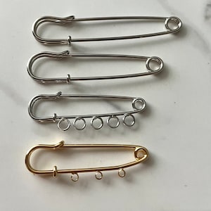May include: Four safety pins, two silver and two gold, with jump rings attached to the bottom of the two gold pins.