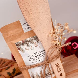 May include: A festive gift set comprising a small wooden spatula and a brown paper bag, tied with twine. The bag has a label with text and a Christmas tree design. The spatula is engraved with a name and a heart.
