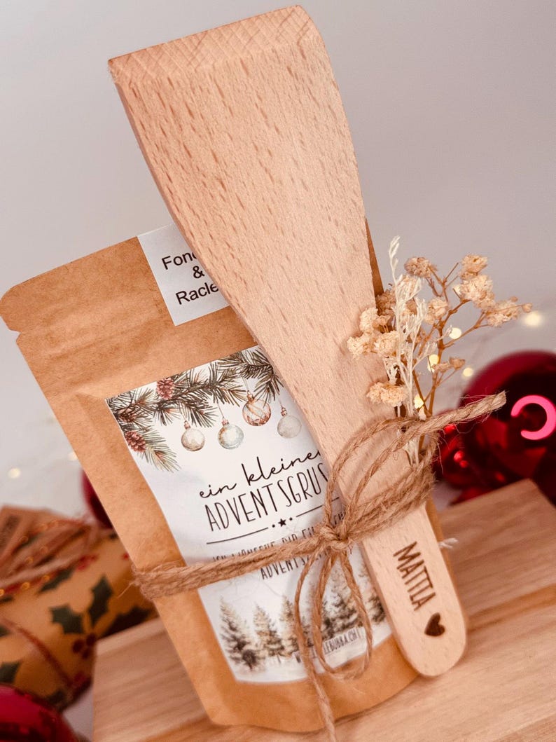 May include: A festive gift set comprising a small wooden spatula and a brown paper bag, tied with twine. The bag has a label with text and a Christmas tree design. The spatula is engraved with a name and a heart.