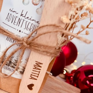 May include: A small gift set featuring a wooden spoon engraved with the name "MATTIA" and a heart symbol. The set is tied with twine and includes a package with text and Christmas ornaments. Red baubles are in the background.