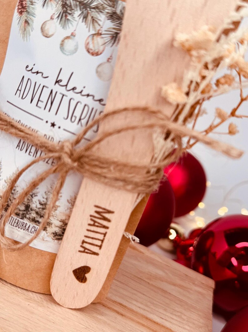 May include: A small gift set featuring a wooden spoon engraved with the name "MATTIA" and a heart symbol. The set is tied with twine and includes a package with text and Christmas ornaments. Red baubles are in the background.