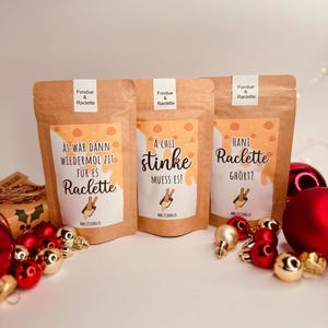 May include: Three brown paper bags labelled "Fondue & Raclette" with Swiss German text and illustrations. The bags are decorated with red and gold Christmas baubles. The text on the bags includes phrases like "stinke" and "Raclette".